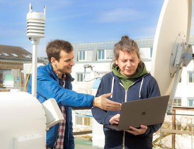 Zwei Männer stehen hinter dem Mikroregenradar und schauen auf einen Laptop, den der eine Mann in der Hand hält. Foto: Swen Reichhold 