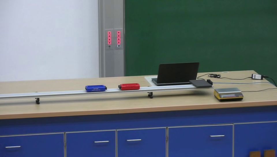 zur Vergrößerungsansicht des Bildes: Ausschnitt eines Experiments mit Rollenfahrbahn auf einem Experimentiertisch auf der zwei Wagen fahren, mit Tafel und Messrechner im Hintergrund, Videoausschnitt: Frank Stallmach, Universität Leipzig