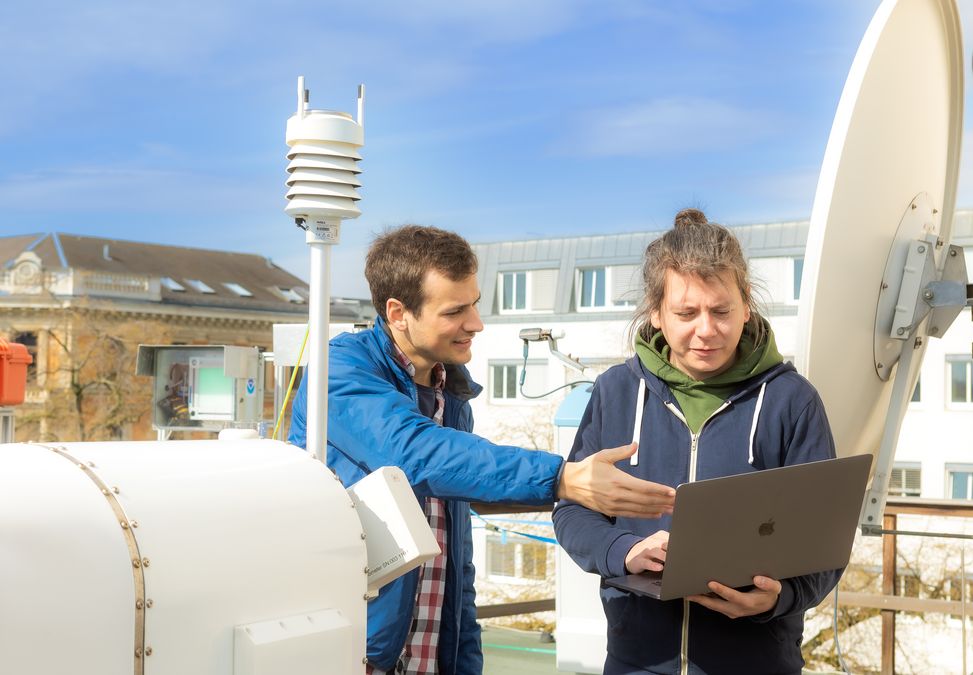 zur Vergrößerungsansicht des Bildes: Zwei Männer stehen hinter dem Mikroregenradar und schauen auf einen Laptop, den der eine Mann in der Hand hält. Foto: Swen Reichhold 