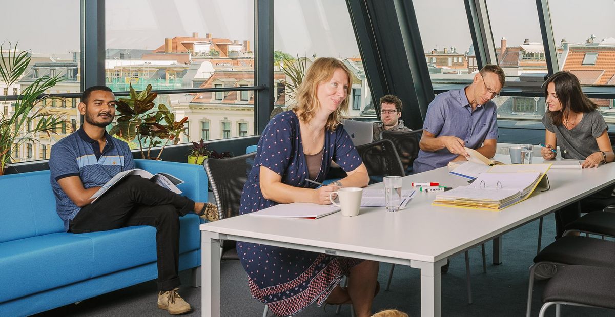 zur Vergrößerungsansicht des Bildes: Die Mitarbeiterinnen und Mitarbeiter der Arbeitsgruppe sitzen im Foyer am Tische und arbeiten. Foto: Stephan Flad