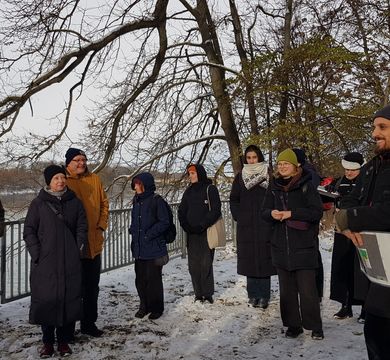 Mehrere Personen stehen im Schnee um einen sprechenden Mann herum, im Hintergrund ein Fluss