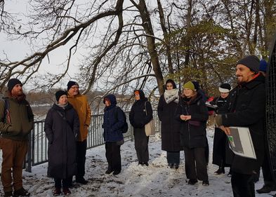 Mehrere Personen stehen im Schnee um einen sprechenden Mann herum, im Hintergrund ein Fluss