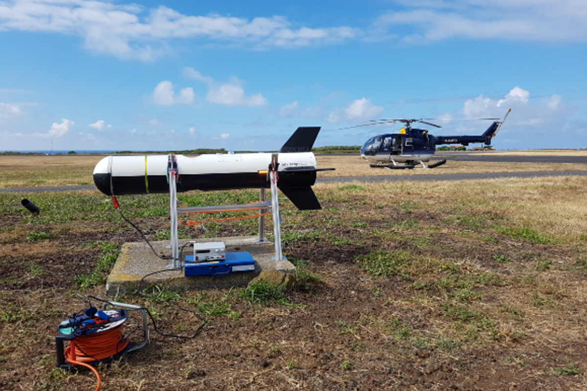 zur Vergrößerungsansicht des Bildes: Das Bild zeigt die Messplattform SMART-HELIOS mit der Form eines Torpedos die am Boden steht und bei der Messung am Hubschrauber hängt. Foto: Felix Lauermann