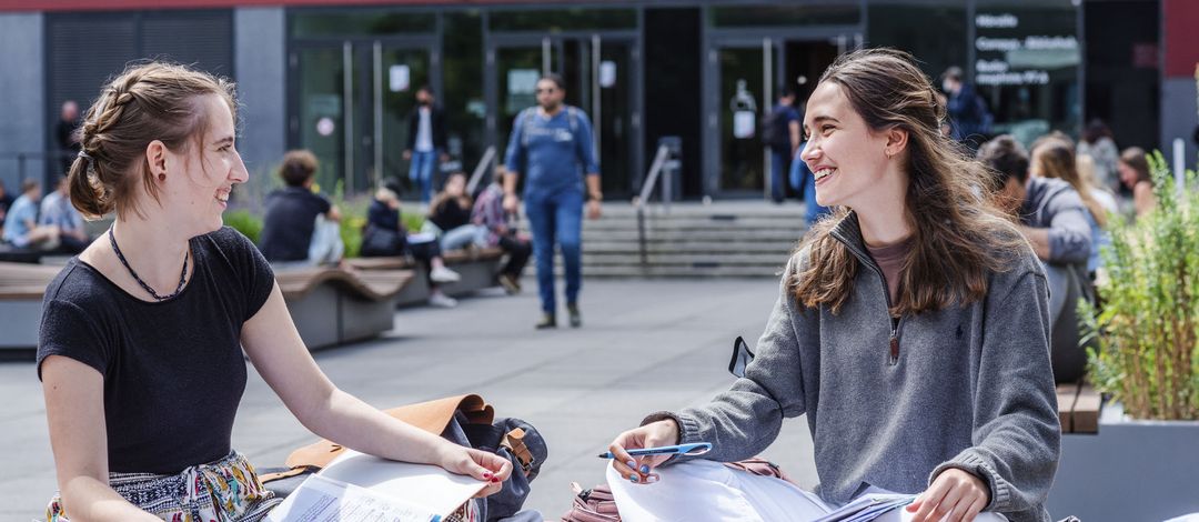 Zwei Studentinnen sitzen auf dem Uni-Innenhof im Schneidersitz auf einer Bank und unterhalten sich. Im Hintergrund sind unscharf Menschen zu sehen, die ins Hörsaalgebäude gehen.